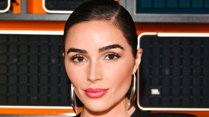 Olivia Culpo smiles for the camera sporting a pink glossy lip, slicked-back hairdo and oversized silver earrings.